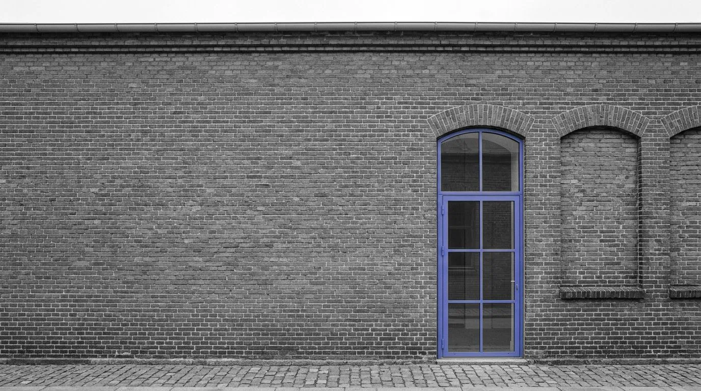 An old brick industrial wall with a long plain stretch, one new steel-framed glass door in purple and bricked-up arched windows beside it